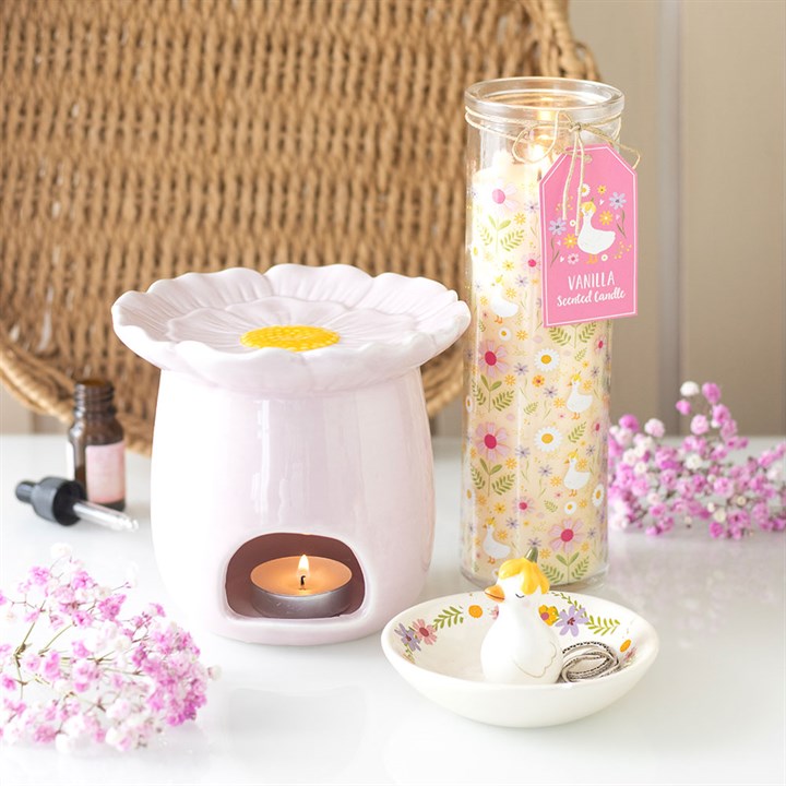 White wax warmer with a lit candle, jar of vanilla-scented candle, and small duck-shaped container on a table with flowers.