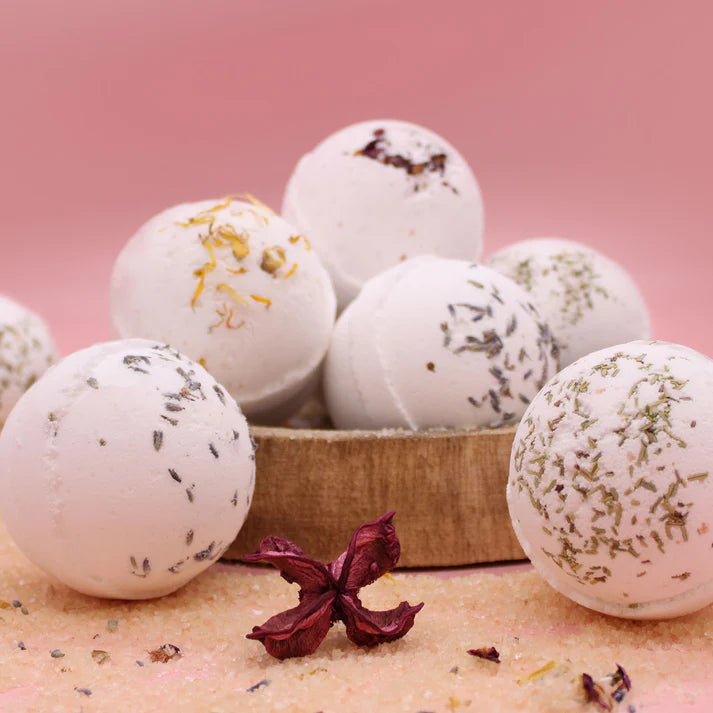 An assortment of bath bombs with various toppings and embedded herbs, displayed in a wooden box against a pink background.