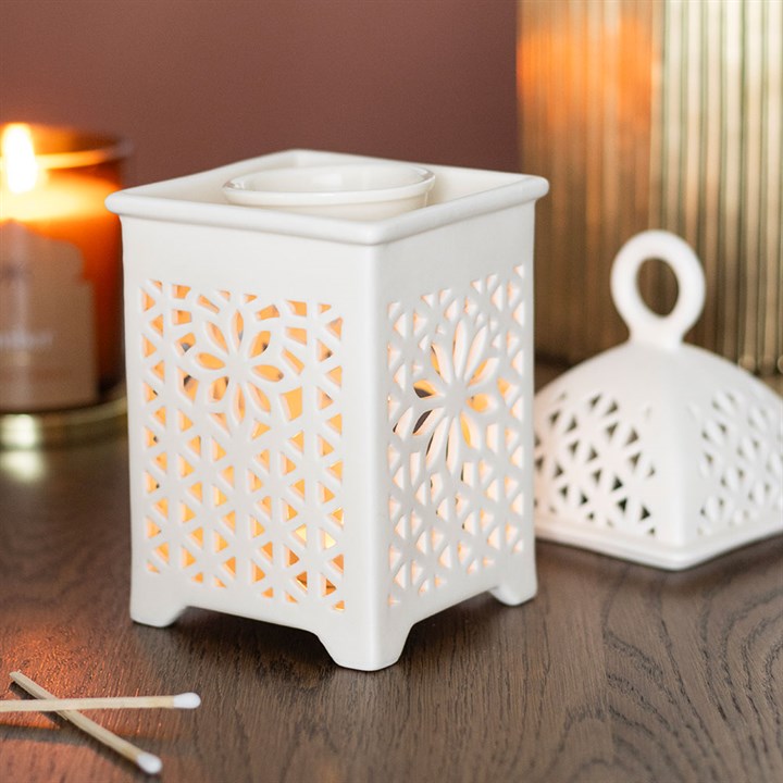 White wax warmer with decorative cut-out design on a wooden surface, with a lit candle in the background.