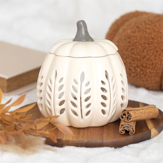 White pumpkin-shaped lantern with cut-out patterns on a wooden surface with autumn leaves and cinnamon sticks.
