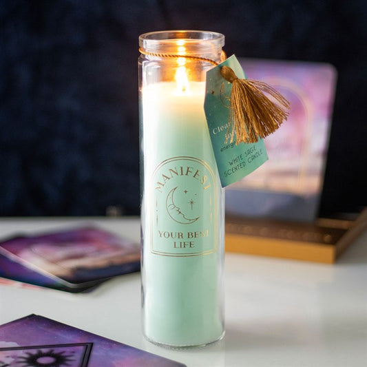 A white tube candle with clear quartz crystal chips inside, placed in a clear glass holder. The candle is lit, creating a warm glow, and has a label with the text 'MANIFEST YOUR BEST LIFE'.