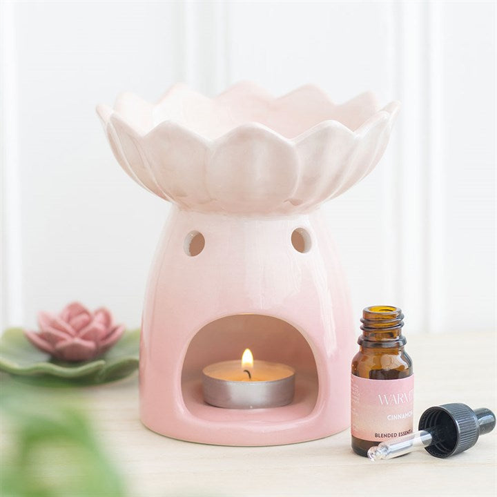 A pink water lily ceramic oil burner with a lit tealight candle inside, accompanied by a small bottle of fragrance oil and a dropper, against a soft-focus background.