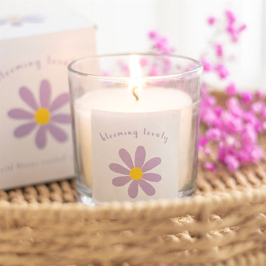 A white candle with a floral design, labeled 'Blooming Lovely', placed on a woven surface, with a box and purple flowers in the background.