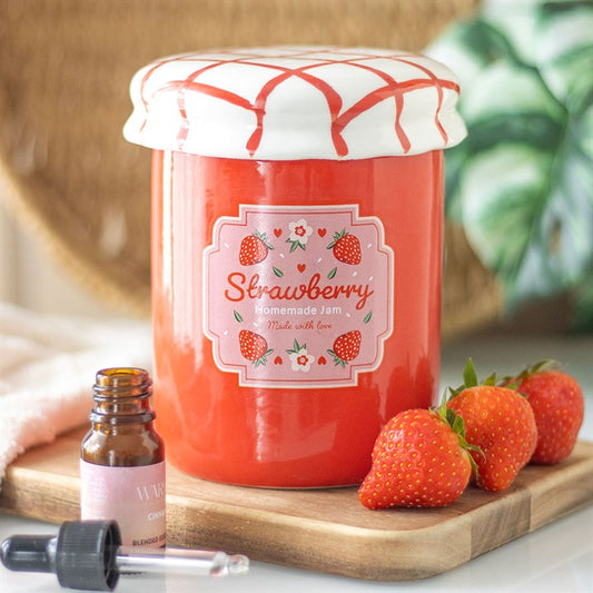 A red vintage-style jam jar oil burner and wax warmer with a white lid, labeled 'Strawberry Jam', accompanied by a small dropper bottle and fresh strawberries.