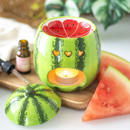 A ceramic oil burner and wax warmer designed to look like a watermelon with a green rind, red interior, and decorative facial expression. A tealight candle is lit inside it, and watermelon slices are placed around it.