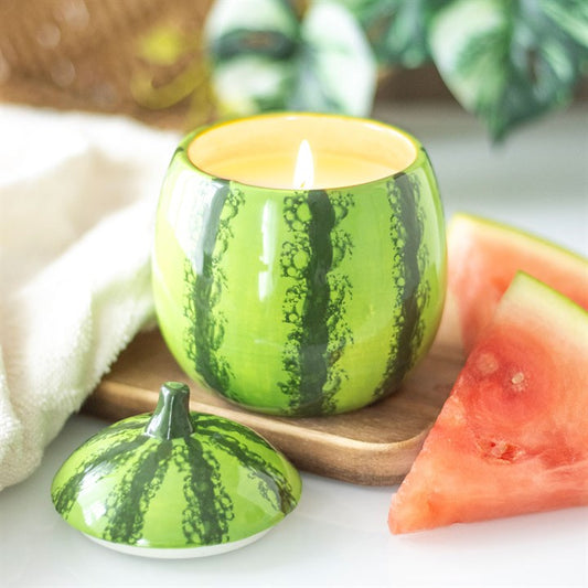 A green candle jar designed to look like a watermelon with a slice of watermelon next to it, placed on a wooden surface.