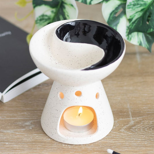 A white ceramic oil burner and wax warmer with a split yin and yang design on the top dish, sitting on a wooden surface with a lit candle below.