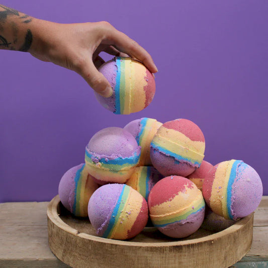 A person's hand holding a multicolored bath bomb with a stack of colorful bath bombs in a wooden bowl on a purple background.