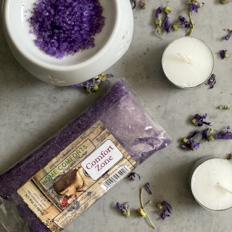 A variety of home fragrance products including wax melts, simmering granules, and a room spray displayed with purple flower petals and candles.