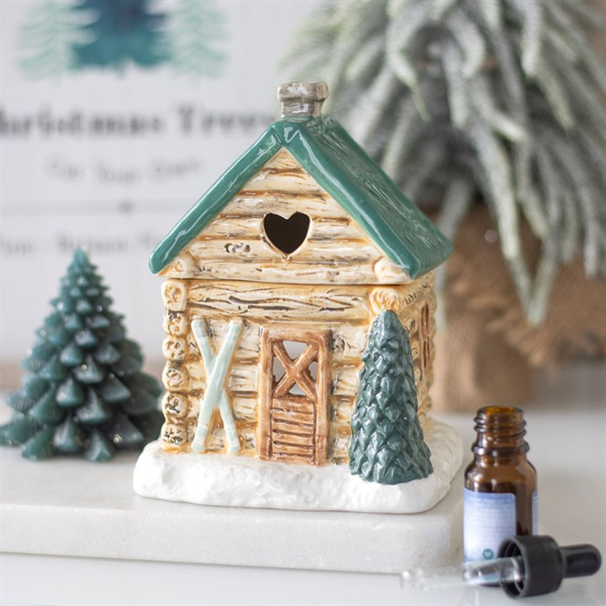 Decorative log cabin with a heart-shaped window, surrounded by small trees and a bottle of essential oil.