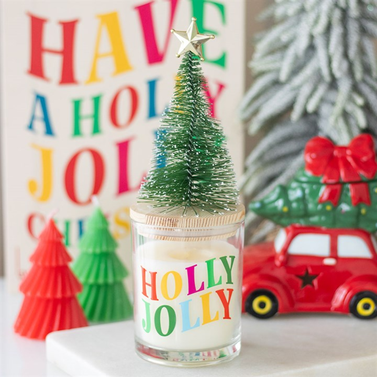 Candle with 'Holly Jolly' text and small Christmas tree decoration in front of colorful lettering.