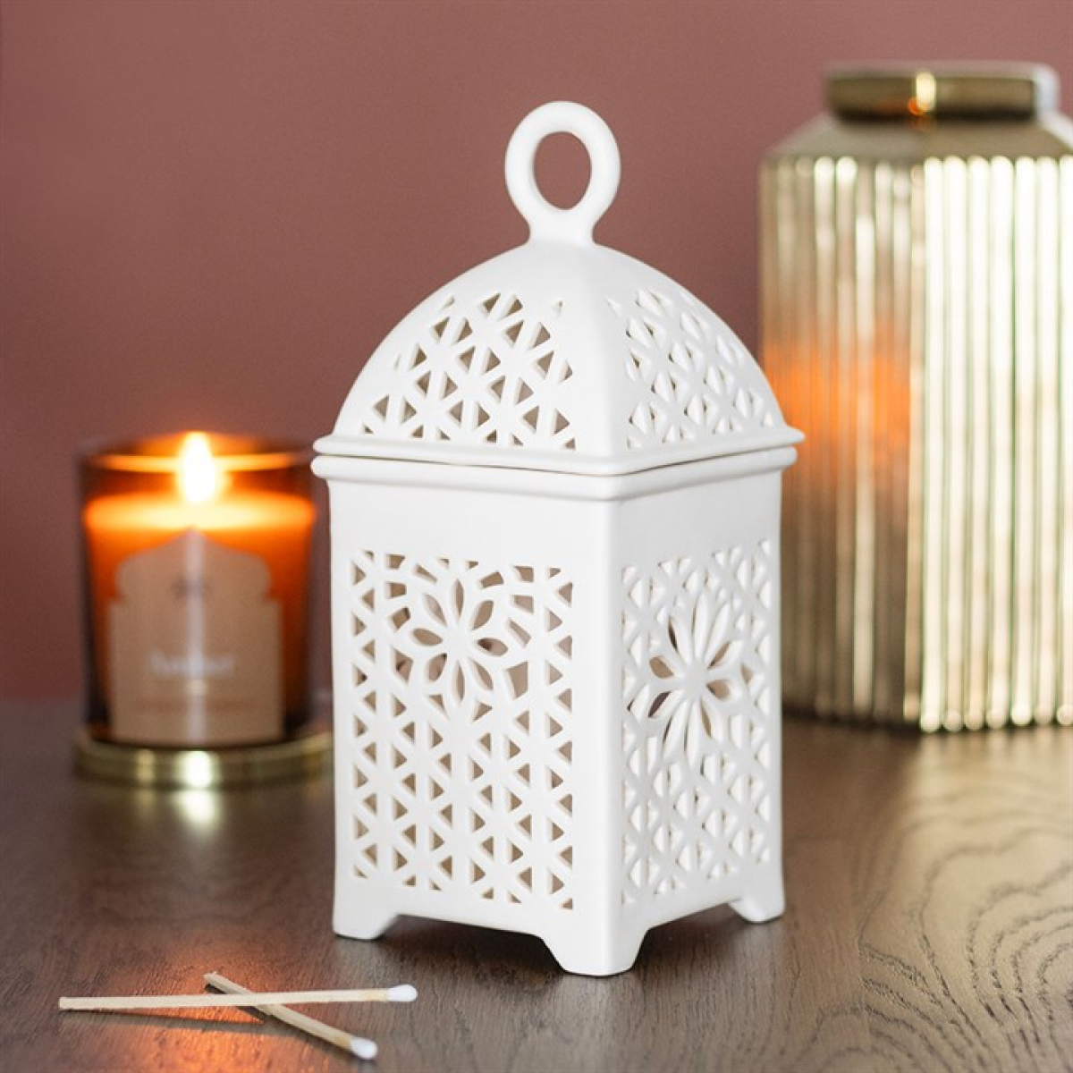 White decorative lantern with intricate patterns on a wooden surface, with a lit candle and another lantern in the background.
