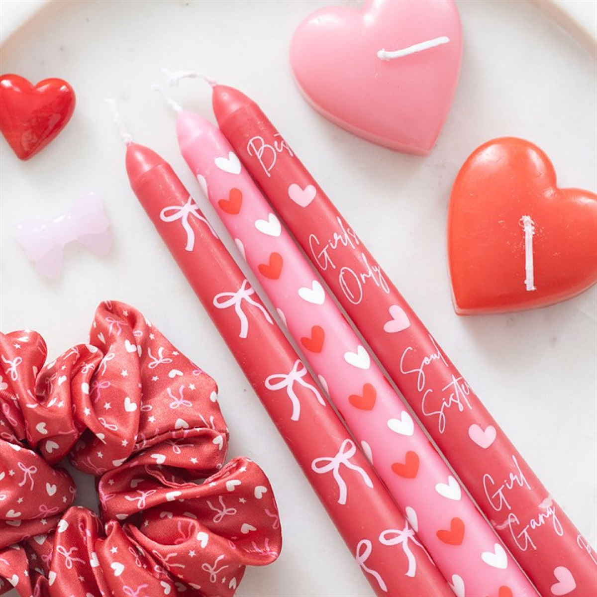 Two pink markers with heart and bow designs, a red scrunchie, and heart-shaped candles on a white surface.