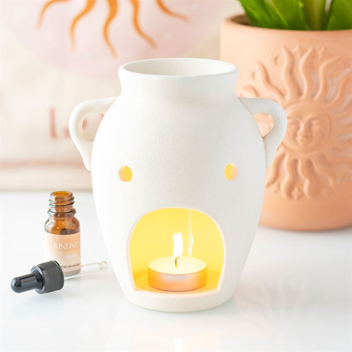 White ceramic diffuser with a lit candle inside, surrounded by a bottle of essential oil and a plant.