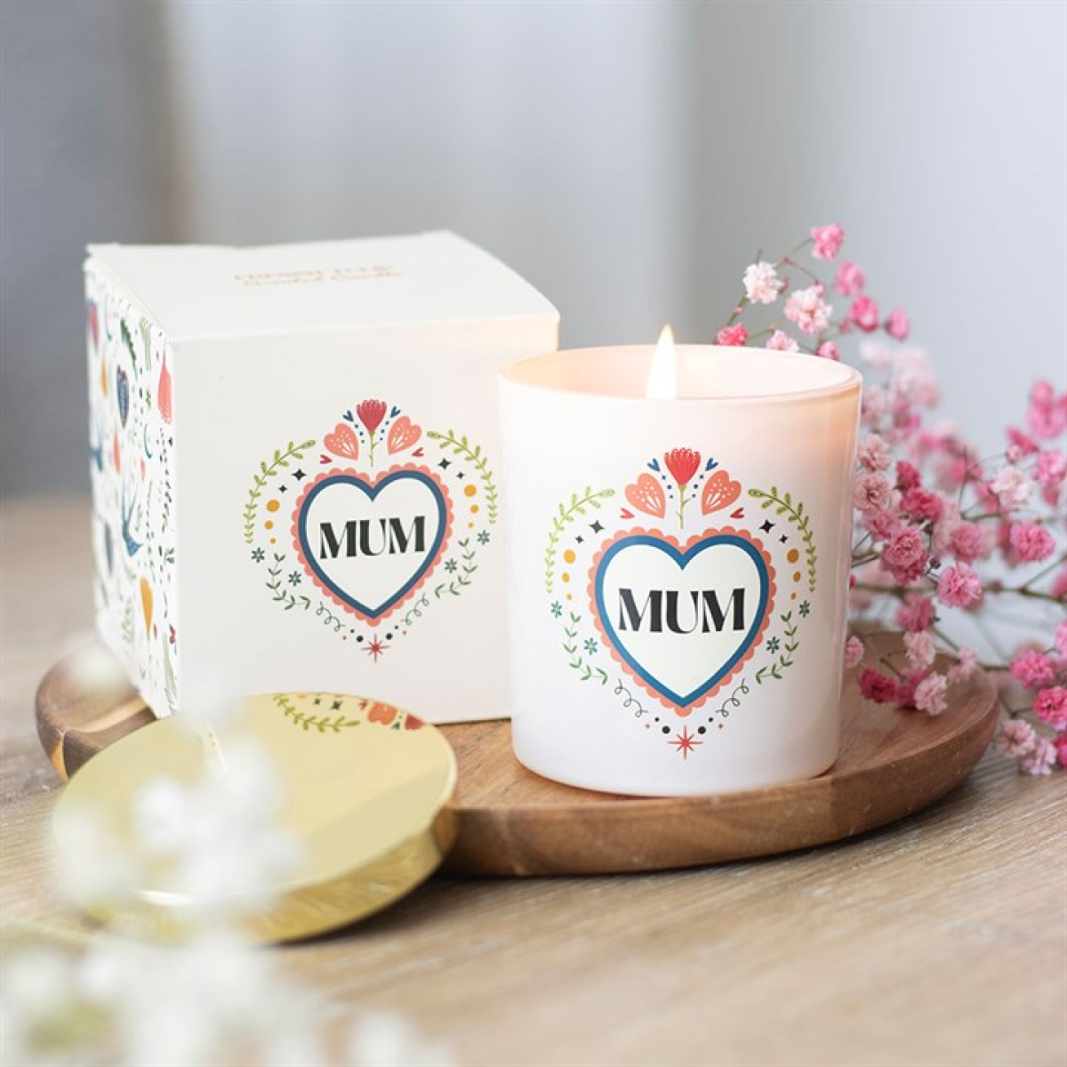 Candle with 'MUM' heart design on a wooden tray with flowers in the background
