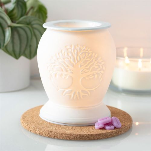A white ceramic electric wax warmer with a Tree of Life design, illuminated by a warm yellow glow, placed on a cork mat.